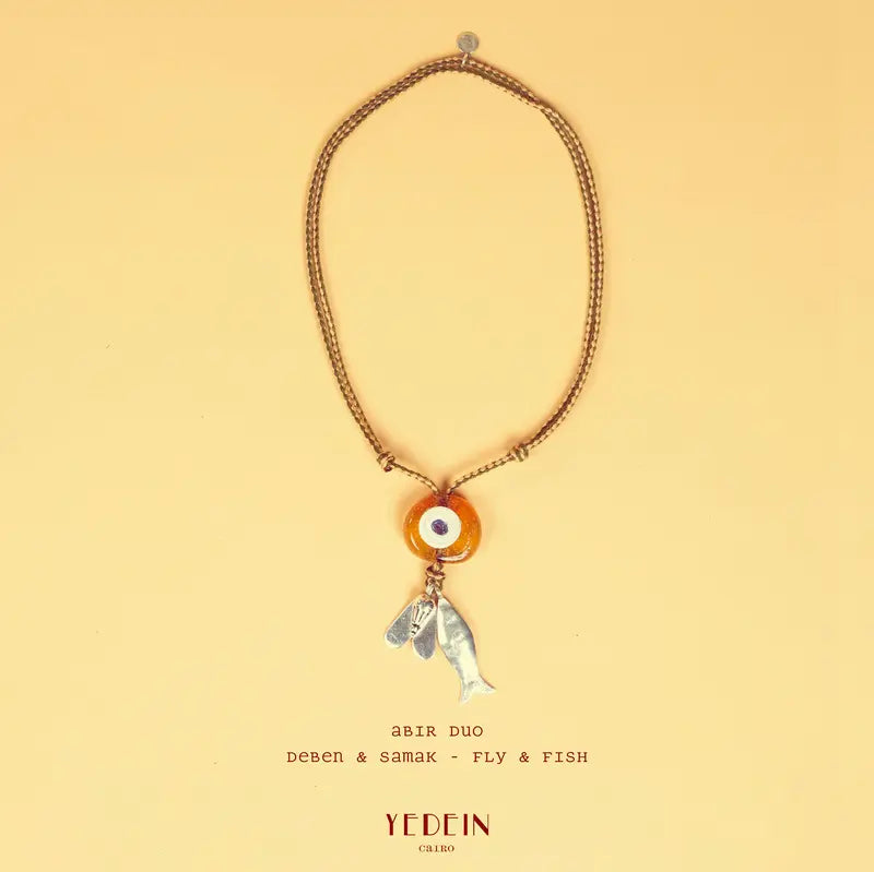 Collier Abir Duo avec un œil ambre et deux charms Samak et Deben sur fond beige